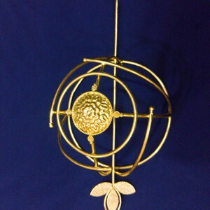 May include: A gold-coloured, ornate pendant with a floral design at the centre. The pendant features a spherical cage-like structure with a vertical rod and a three-leaf design at the bottom. The background is a solid dark blue.