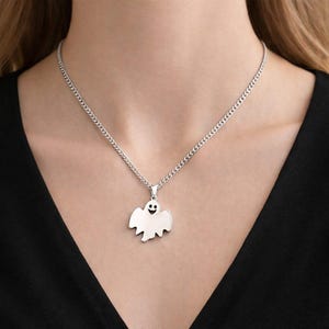 Puede incluir: Un collar plateado con un colgante en forma de fantasma sonriente. El colgante cuelga de una cadena fina. El collar se muestra sobre una blusa negra.