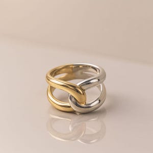 May include: A two-tone ring with a linked design. The ring features a gold-colored section intertwined with a silver-colored section. The ring is displayed on a neutral background, highlighting its contrasting colors and unique shape.