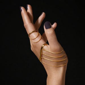 May include: Gold-toned hand jewelry set. The set includes a ring with two connected bands and a hand chain with multiple delicate strands. The jewelry is displayed against a dark background.