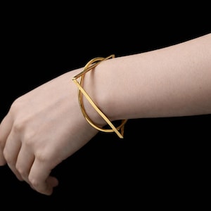May include: A gold-coloured bracelet with a geometric design. The bracelet features a square and curved shape, and is worn on a wrist against a black background. The jewellery is made of metal.