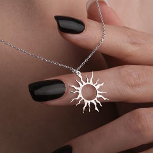 Puede incluir: Un collar plateado con un colgante de sol. El colgante de sol tiene un centro circular con rayos que se extienden hacia afuera. La cadena del collar es delicada y parece ser de plata. El fondo está borroso.