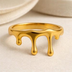 May include: A gold-colored ring with a unique design. The ring features a band with a dripping effect, creating a fluid, melted appearance. The ring is made of a shiny, reflective metal, and the design is eye-catching and modern.