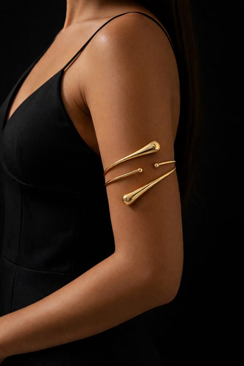 May include: Gold-toned arm cuff with a minimalist design. The jewelry features three curved bands with teardrop-shaped accents. The arm cuff is worn on the upper arm, against a dark background. The accessory is a stylish piece of body jewelry.
