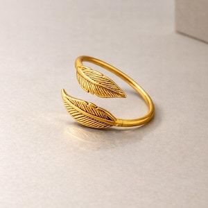Puede incluir: Un brazalete dorado con dos diseños detallados de plumas. El brazalete tiene un diseño abierto, con las plumas colocadas en cada extremo. La joya está hecha de un material metálico brillante.