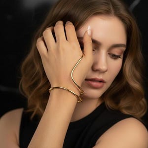 Può includere: Bracciale da mano e bracciale a polsino dorati. Il bracciale da mano si curva sul dorso della mano, collegandosi al bracciale a polsino sul polso. I gioielli sono in metallo lucido e riflettente. Lo sfondo è nero.