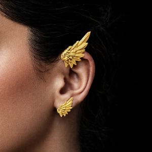 Könnte beinhalten: Goldfarbenes Ohr-Manschetten- und Ohrring-Set in Flügelform. Die Ohr-Manschette umschließt das obere Ohr, während der Ohrring ein kleineres Flügeldesign ist. Der Schmuck ist detailliert mit einem strukturierten Federmuster vor dunklem Hintergrund.