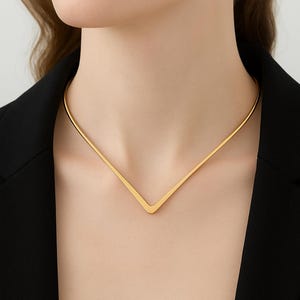 Puede incluir: Un collar dorado con un colgante en forma de V. El collar lo lleva una persona con una blazer negra. El colgante tiene un diseño sencillo y moderno, y el collar parece ser de metal.