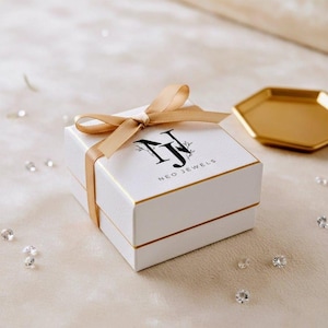 May include: A white jewelry box with gold trim and a matching ribbon. The box has the words "NEO JEWELS" printed on the top. A gold-colored hexagonal tray is in the background.