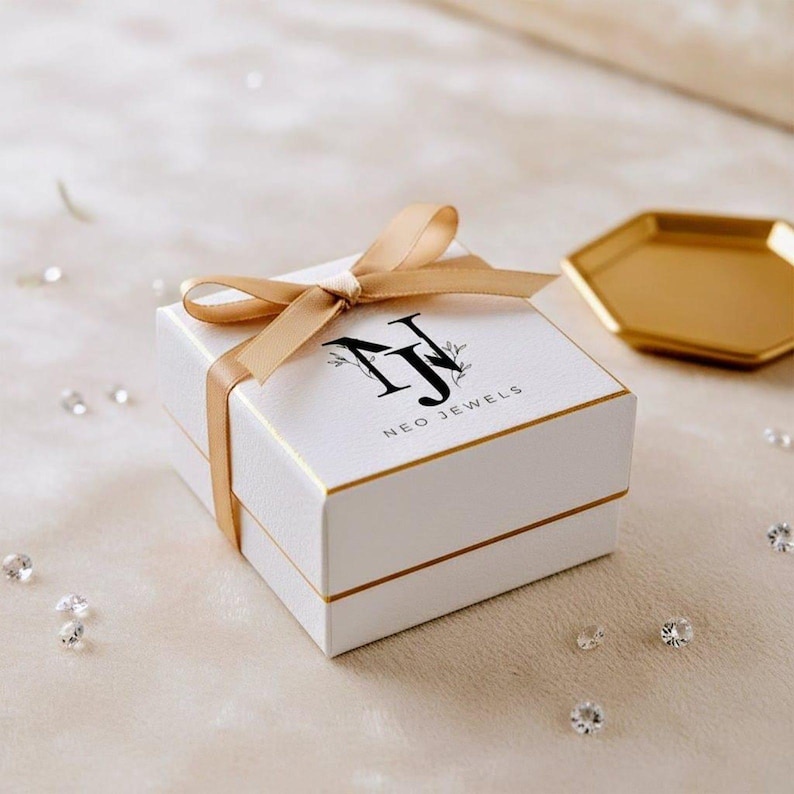 May include: A white jewelry box with gold trim and a matching ribbon. The box has the words "NEO JEWELS" printed on the top. A gold-colored hexagonal tray is in the background.