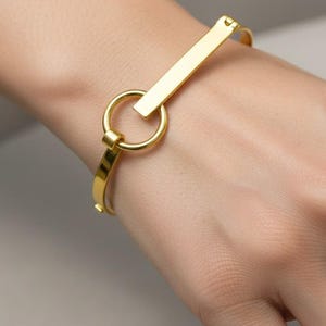 May include: A gold-coloured bangle bracelet featuring a rectangular bar and a circular ring. The bracelet is worn on a wrist, highlighting its simple, elegant design. The polished metal reflects light, creating a subtle shine.