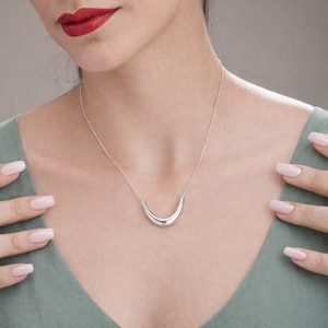 Puede incluir: Un collar plateado con un colgante curvo en forma de media luna. El collar se lleva en una fina cadena. La persona lleva una blusa verde y tiene pintalabios rojo. Un diseño sencillo y elegante.