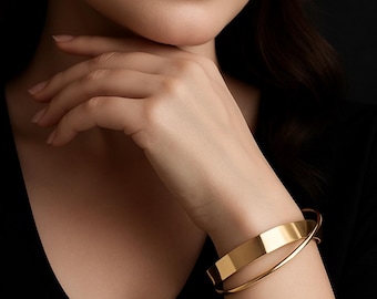 May include: A gold-colored bracelet with a flat, rectangular design and a curved band. The jewelry is worn on the wrist. The image is set against a dark background.