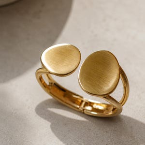 Può includere: Un bracciale rigido dorato con cerniera, caratterizzato da due dischi piatti e circolari sull'apertura. Il metallo presenta una finitura spazzolata e opaca, e la struttura è composta da due fili paralleli uniti da una cerniera sul retro.