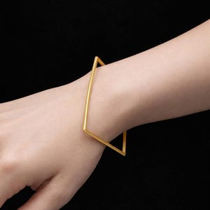 May include: A gold-coloured square bangle bracelet. The bracelet has a simple, geometric design, with a square shape. The jewellery is worn on a wrist against a black background. The bracelet is made of metal.
