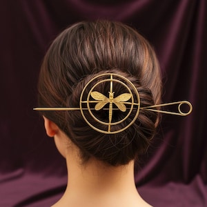 Puede incluir: Horquilla dorada con diseño de libélula. La horquilla tiene un marco circular con una libélula en el centro y un alfiler largo y recto que se extiende desde un lado. La libélula tiene alas y cuerpo texturizados.