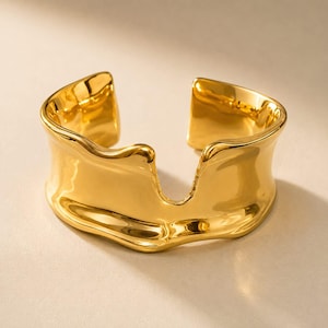 May include: A wide, polished gold-tone cuff bracelet featuring an irregular, organic, and wavy silhouette. The metal surface is smooth and reflective, with a prominent U-shaped cutout at the centre of the front, creating an open-ended design.