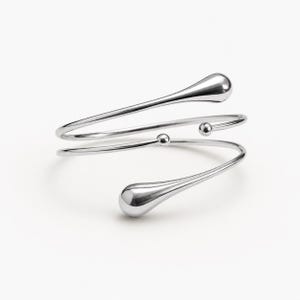 May include: A silver-toned arm cuff with a minimalist design. The cuff features a spiral shape with two teardrop-shaped ends and small spherical accents. The polished metal reflects light, creating a sleek and modern aesthetic.