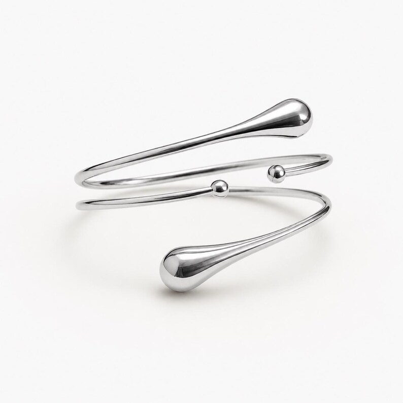 May include: A silver-toned arm cuff with a minimalist design. The cuff features a spiral shape with two teardrop-shaped ends and small spherical accents. The polished metal reflects light, creating a sleek and modern aesthetic.