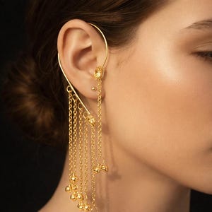 May include: A gold ear cuff with a detailed design. The cuff wraps around the ear and has hanging chains and small decorative features. The jewellery is shown against a dark backdrop.