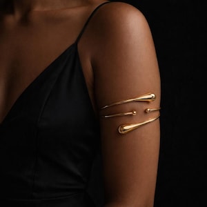 May include: Gold-toned arm cuff with a minimalist design. The cuff features three curved bands, with teardrop-shaped ends and a small sphere accent. The jewelry is worn on the upper arm against a black background.
