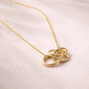 Könnte beinhalten: Eine goldfarbene Halskette mit einem Anhänger in Form eines verschlungenen Knotens. Die Kette ist filigran und wird auf einem weichen, hellen Stoff präsentiert.