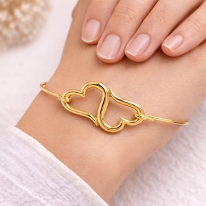 Könnte beinhalten: Ein goldfarbenes Armband mit zwei verbundenen herzförmigen Anhängern. Das Armband ist ein zartes, dünnes Band, und die Herzen sind glatt geformt. Das Armband wird an einem Handgelenk getragen, vor einem weißen Hemd.