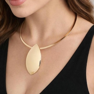Pode incluir: Um colar dourado com um design minimalista. Possui uma gola rígida e curva e um grande pingente em forma de gota. O pingente e a gola têm uma superfície polida e reflexiva. O colar é usado sobre um top preto.
