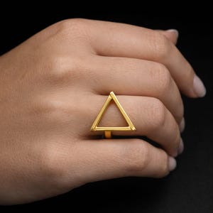 May include: A gold-coloured ring with a geometric triangle design. The ring is worn on a finger, highlighting its minimalist and modern aesthetic. The triangle is open, creating a unique and stylish look.