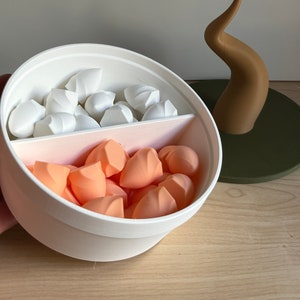 May include: A white and pink cosmetic sponge holder with a divider, filled with white and peach-colored makeup sponges. A brown decorative element sits on a green circular base in the background.