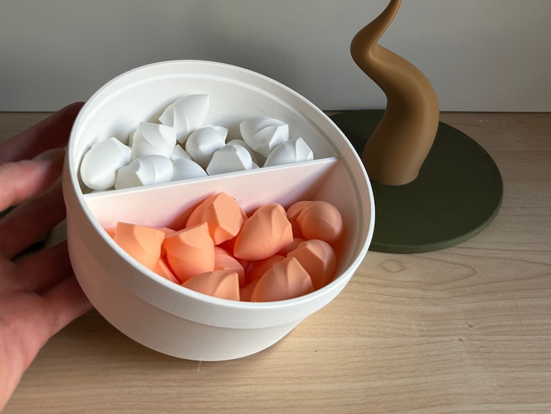 May include: A white and pink cosmetic sponge holder with a divider, filled with white and peach-colored makeup sponges. A brown decorative element sits on a green circular base in the background.