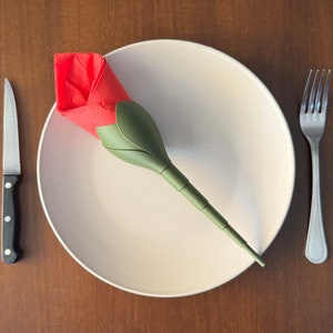 May include: A white plate with a red and green napkin folded into the shape of a rose. A silver fork and knife are placed on either side of the plate.