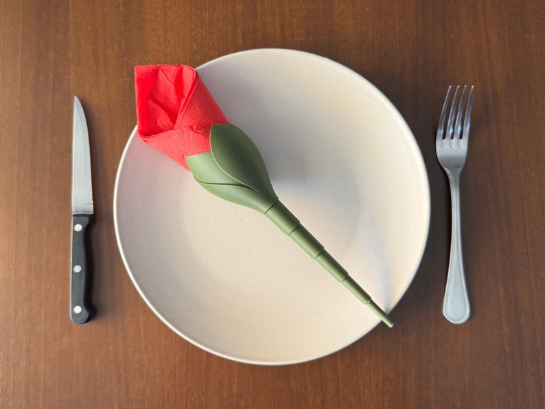 May include: A white plate with a red and green napkin folded into the shape of a rose. A silver fork and knife are placed on either side of the plate.