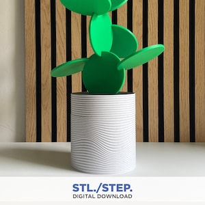 Puede incluir: Una escultura de planta impresa en 3D con hojas verdes y una maceta blanca texturizada. La escultura está delante de una pared de panel de madera con líneas verticales negras.