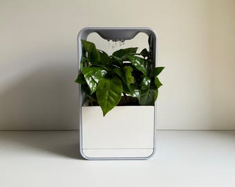 May include: A gray and white indoor plant growing system with a green plant inside. The system has a water reservoir and a light source.
