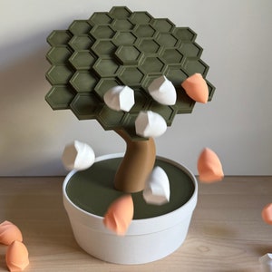 May include: A whimsical, modern bonsai tree sculpture with a green hexagonal canopy and a brown trunk, set in a white pot. White and peach-colored objects appear to be falling from the tree, creating a dynamic visual.