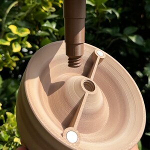 May include: A light brown, wooden-like object with a central screw and a brown stem. The object has a circular base with two arms and two small, white circular magnets. The background is blurred green foliage.