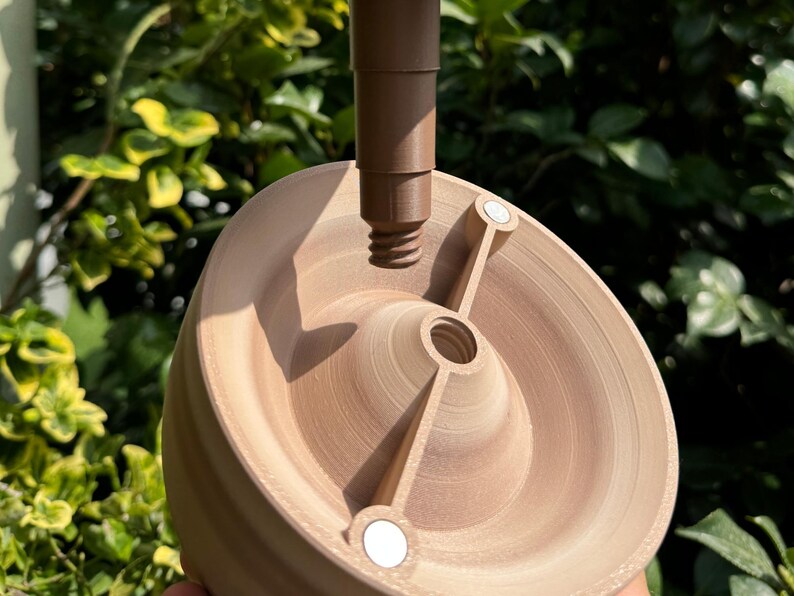 May include: A light brown, wooden-like object with a central screw and a brown stem. The object has a circular base with two arms and two small, white circular magnets. The background is blurred green foliage.