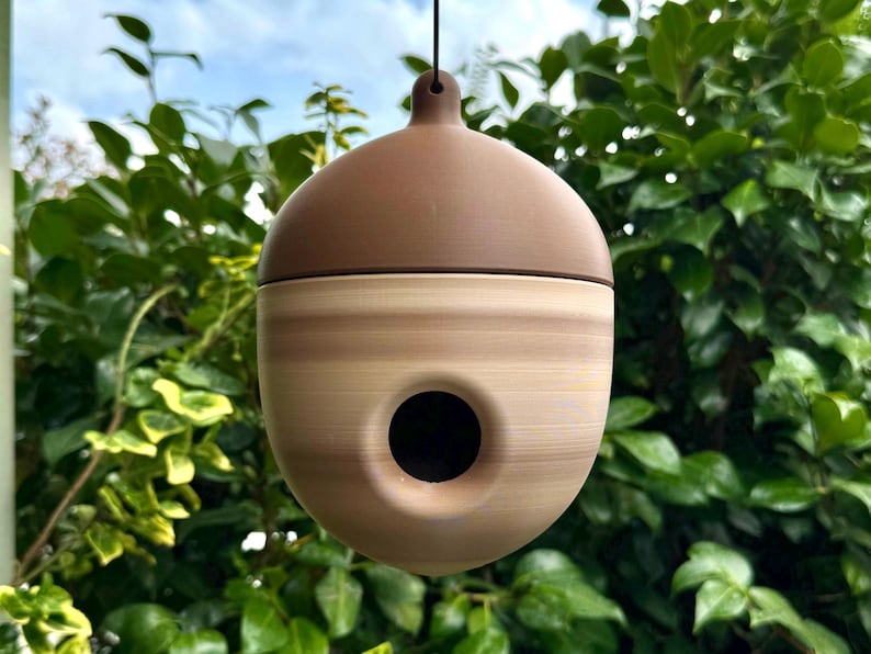 May include: A brown and tan acorn-shaped birdhouse with a circular entry hole. The birdhouse is suspended by a black string against a backdrop of green foliage and a blue sky. The top is a darker brown than the body.