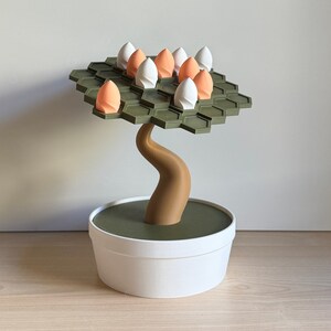 May include: A decorative bonsai tree sculpture with a white pot and a brown trunk. The tree's canopy is a hexagonal grid in olive green, holding white and orange egg-shaped ornaments. The sculpture is a unique home decor item.