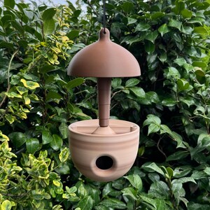 May include: A hanging bird feeder with a domed top and a rounded base. The feeder is made of a light brown material and has a circular opening on the side. The background is filled with green foliage.