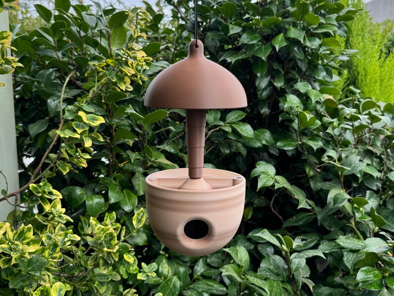 May include: A hanging bird feeder with a domed top and a rounded base. The feeder is made of a light brown material and has a circular opening on the side. The background is filled with green foliage.