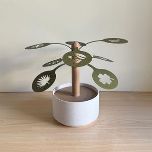 Könnte beinhalten: Ein dekorativer Baum aus goldenem Metall mit einem weißen Keramiksockel. Der Baum hat mehrere Äste mit ausgeschnittenen Blattmotiven. Die Blätter haben verschiedene Formen, darunter ein Kleeblatt, ein Palmblatt und ein Farn.