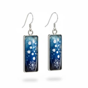 May include: A pair of rectangular drop earrings with a silver frame. The earrings display a blue background with a white floral design. The design includes white dots and a stylized flower. The earrings hang from silver fish hook ear wires.