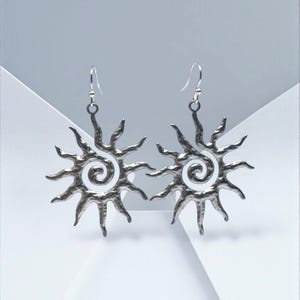 May include: A pair of silver-toned earrings featuring a sunburst design with a central spiral and radiating pointed rays. The metal surface has a hammered, textured finish. Each earring hangs from a simple silver-toned hook wire for attachment.
