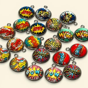 May include: A selection of circular charms with comic book-style designs and silver-coloured metal edges. Each charm displays a different action word in bold, colourful text, including "ZAP!", "POW!", and "BOING!". The charms are arranged on a light-coloured surface.