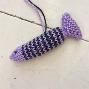 Può includere: Un oggetto lavorato a maglia a mano nei toni del viola e del blu navy, con un motivo strutturato. L'oggetto ha una forma arrotondata con una piccola maniglia rettangolare. Due fili di filo, uno viola e uno blu scuro, si estendono dalla parte superiore.