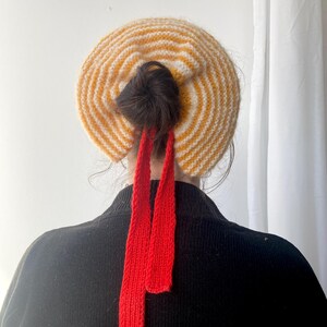 Puede incluir: Un gorro de punto con un patrón de rayas amarillas y blancas. El gorro tiene un moño marrón oscuro y dos largas cintas rojas de punto. La persona lleva un jersey negro.