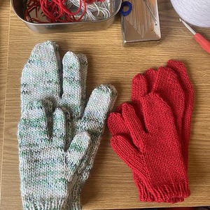 May include: A pair of knitted gloves, one in a speckled green and grey yarn, the other in a solid red. The gloves are handmade and look warm. Knitting supplies, including yarn and needles, are visible in the background.