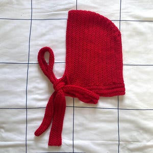 Puede incluir: Un gorro de punto rojo vibrante con lazos largos. El gorro está hecho de una tela de punto texturizada y presenta una parte superior redondeada y un cierre de lazo. Los lazos están anudados en la base del gorro, creando un ajuste seguro. El gorro se muestra sobre una superficie blanca con un patrón de cuadrícula.